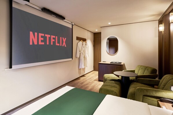 A room with a large screen displaying 'NETFLIX,' a bed, two green armchairs, a small table, and a window with blinds.