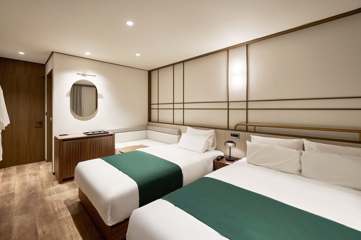 Habitación de hotel con dos camas, un armario de madera, un espejo redondo y una mesita de noche con lámpara.