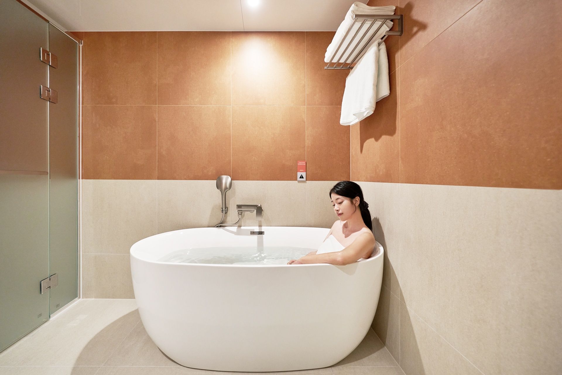 Eine Frau entspannt sich in einer großen Badewanne mit Glasverkleidung, darüber ein Handtuchhalter und eine Wand mit einem Warnschild.