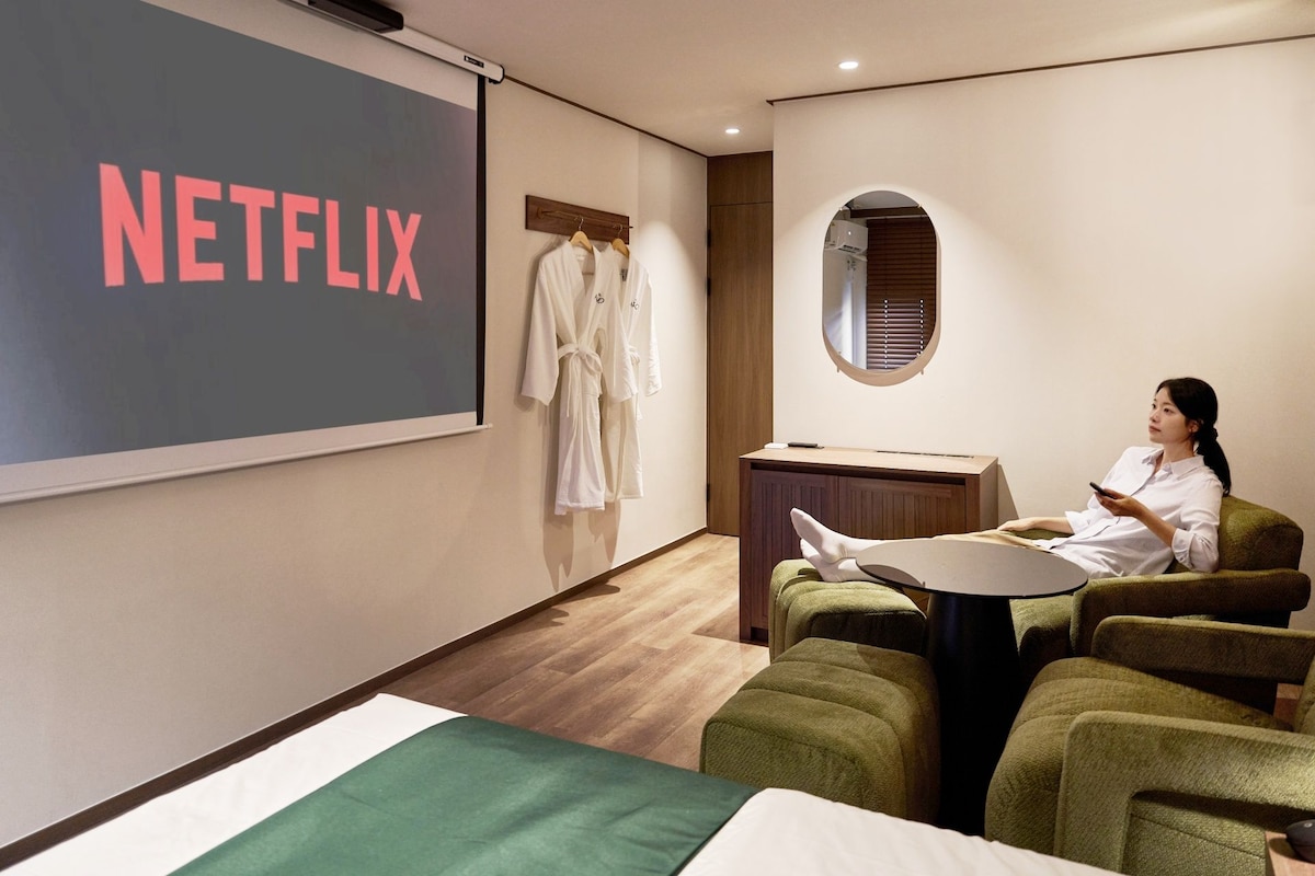 Una habitación con una pantalla grande que muestra 'NETFLIX', un armario de madera, un espejo redondo y una persona sentada en un sofá verde.