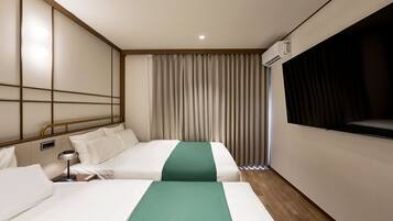 A hotel room with two beds, a bedside table with a lamp, a large window with curtains, and an air conditioning unit.