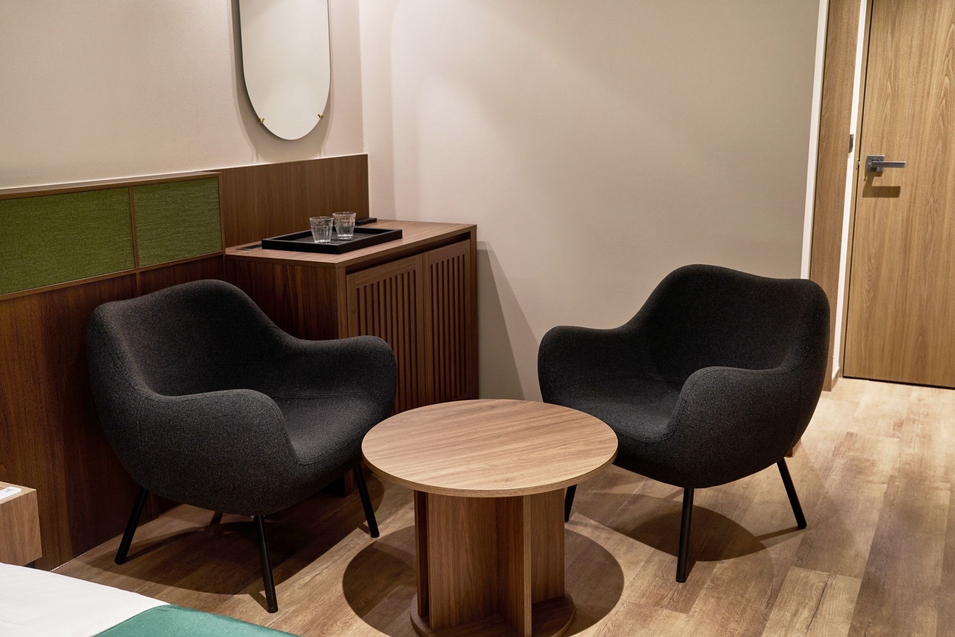 Um quarto com duas poltronas escuras, uma mesa redonda de madeira, um pequeno aparador com copos e uma porta.