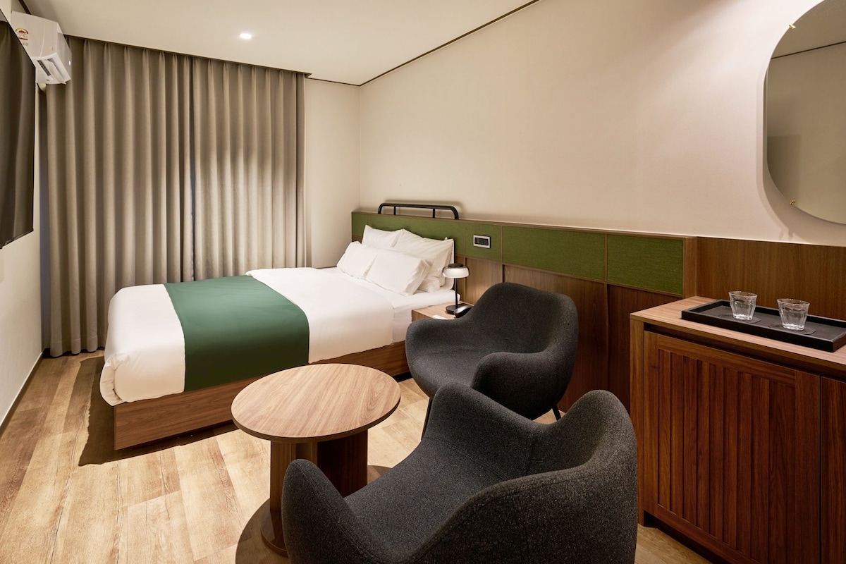 Una habitación de hotel con cama, escritorio, dos sillones, una mesita redonda y un minibar.