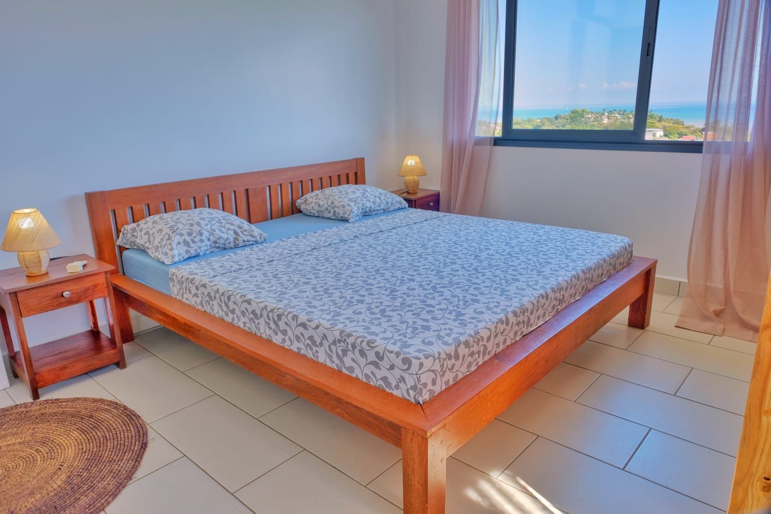 A single bed with a wooden frame and patterned bedding, two bedside lamps, a nightstand, a woven rug, and a view of the sea through a window with sheer curtains.