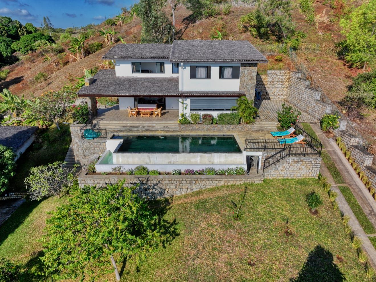 A two-story house with a swimming pool, a patio with outdoor furniture, and a stone staircase leading to the upper level.