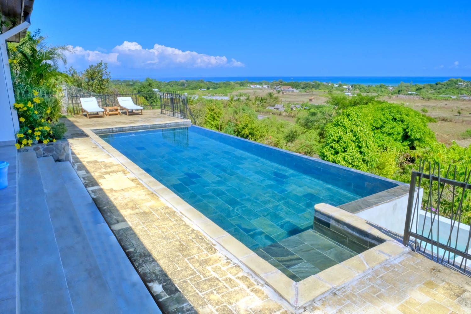 A rectangular swimming pool with a clear blue water surface, surrounded by a stone deck and a metal fence. There are lounge chairs and a small table on the deck.