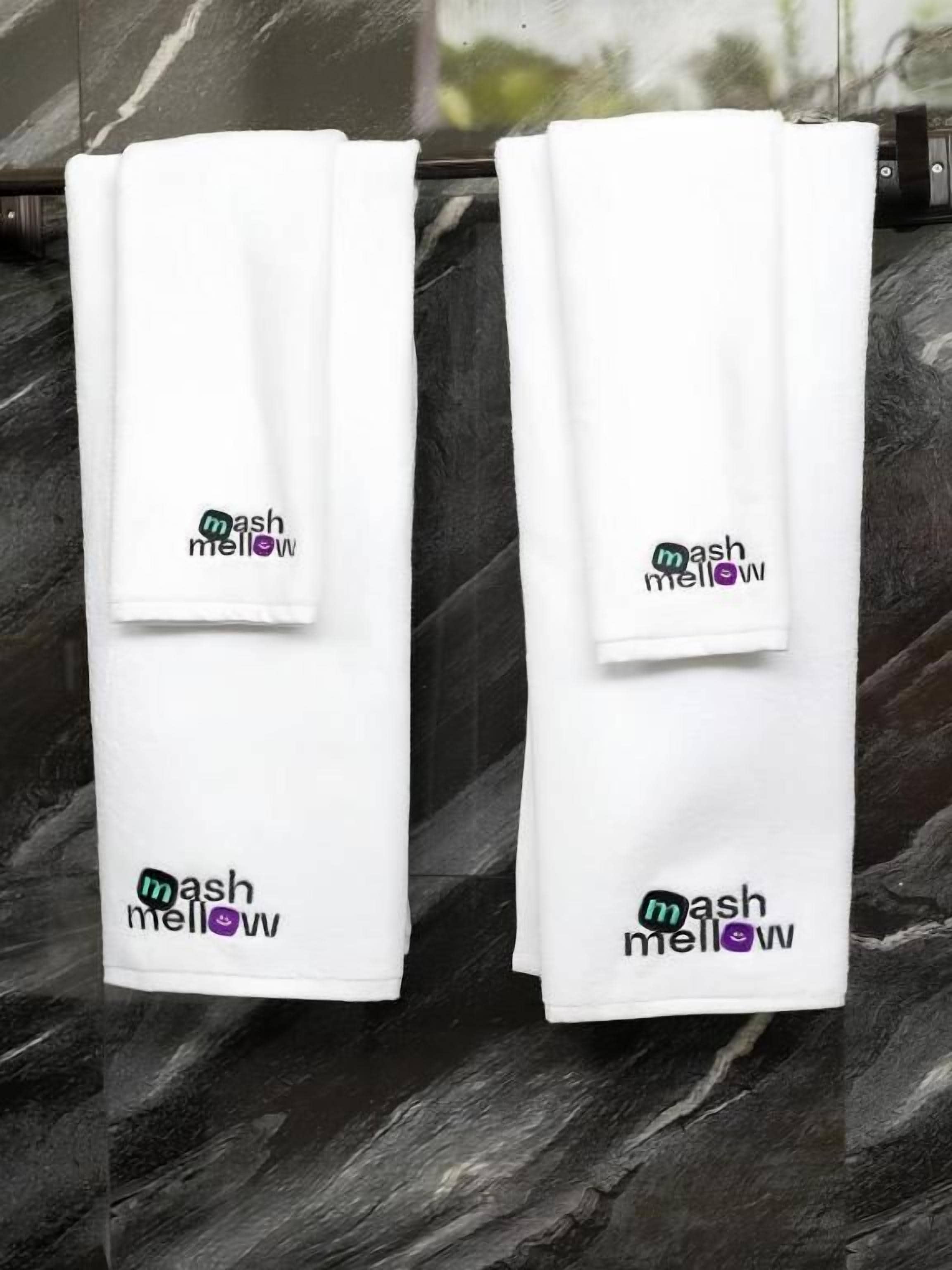 Dos toallas blancas con el logo 'mash mellow' colgadas sobre una superficie oscura y con textura.