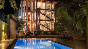 A modern two-story building with a glass facade, a swimming pool in the foreground, and a staircase connecting the two levels.