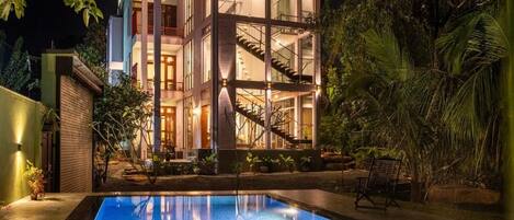 A modern two-story building with a glass facade, a swimming pool in the foreground, and a staircase connecting the two levels.