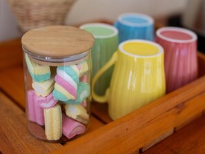 A clear glass jar filled with colorful marshmallows and a wooden lid, placed on a wooden tray with colorful ceramic mugs.