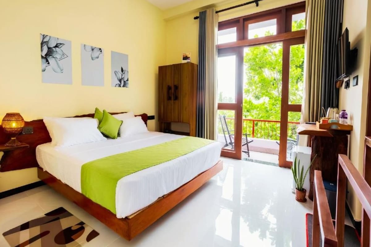 Habitación de hotel con una cama grande, un armario de madera, un escritorio con silla y un balcón con vistas a zonas verdes.