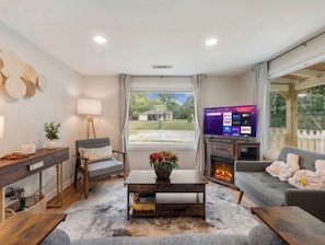 A living room with a fireplace, a television, a sofa, a chair, a coffee table, and a window with a view of a house.