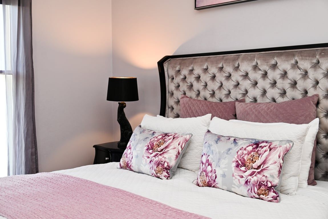 A bedroom with a tufted headboard, a bedside table with a lamp, and floral-patterned pillows.