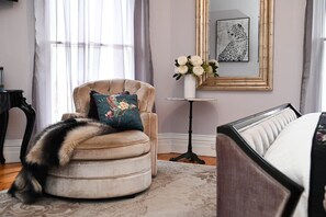 A room with a round armchair, a bed, a small table with a vase of flowers, and a framed artwork on the wall.