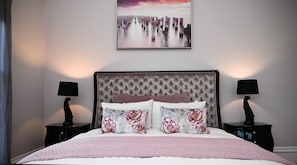 Un dormitorio con cabecera capitonada, almohadas florales, colcha rosa y un cuadro enmarcado sobre la cama.