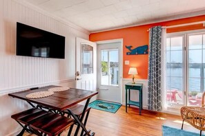 A room with a wooden dining table, a TV mounted on the wall, a door leading outside, and a view of the water.