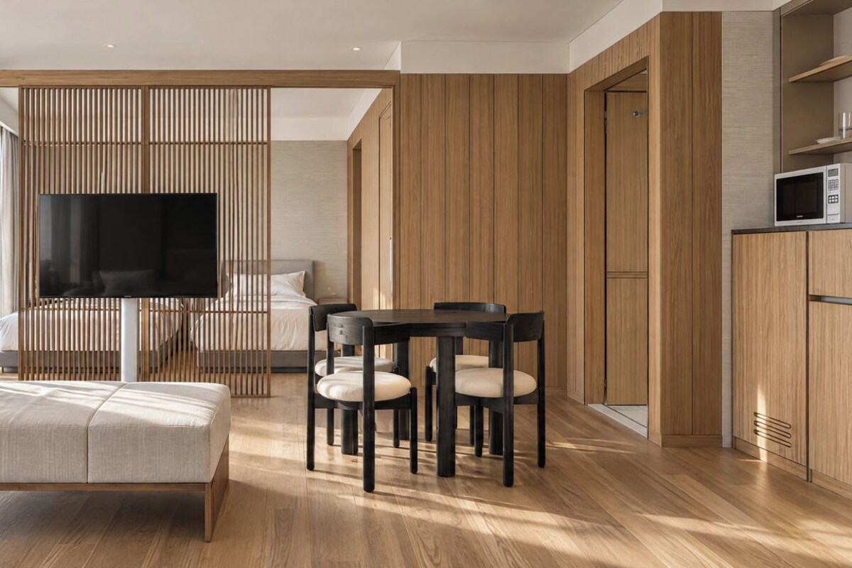 A modern hotel room with a sofa, a dining table with chairs, a TV, and wooden paneling.