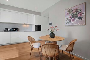 Eine moderne Küche mit weißen Schränken, einem schwarzen Backofen und einer Kaffeemaschine auf der Arbeitsplatte. Ein runder Holztisch mit zwei Korbstühlen und einer Blumenvase steht daneben.
