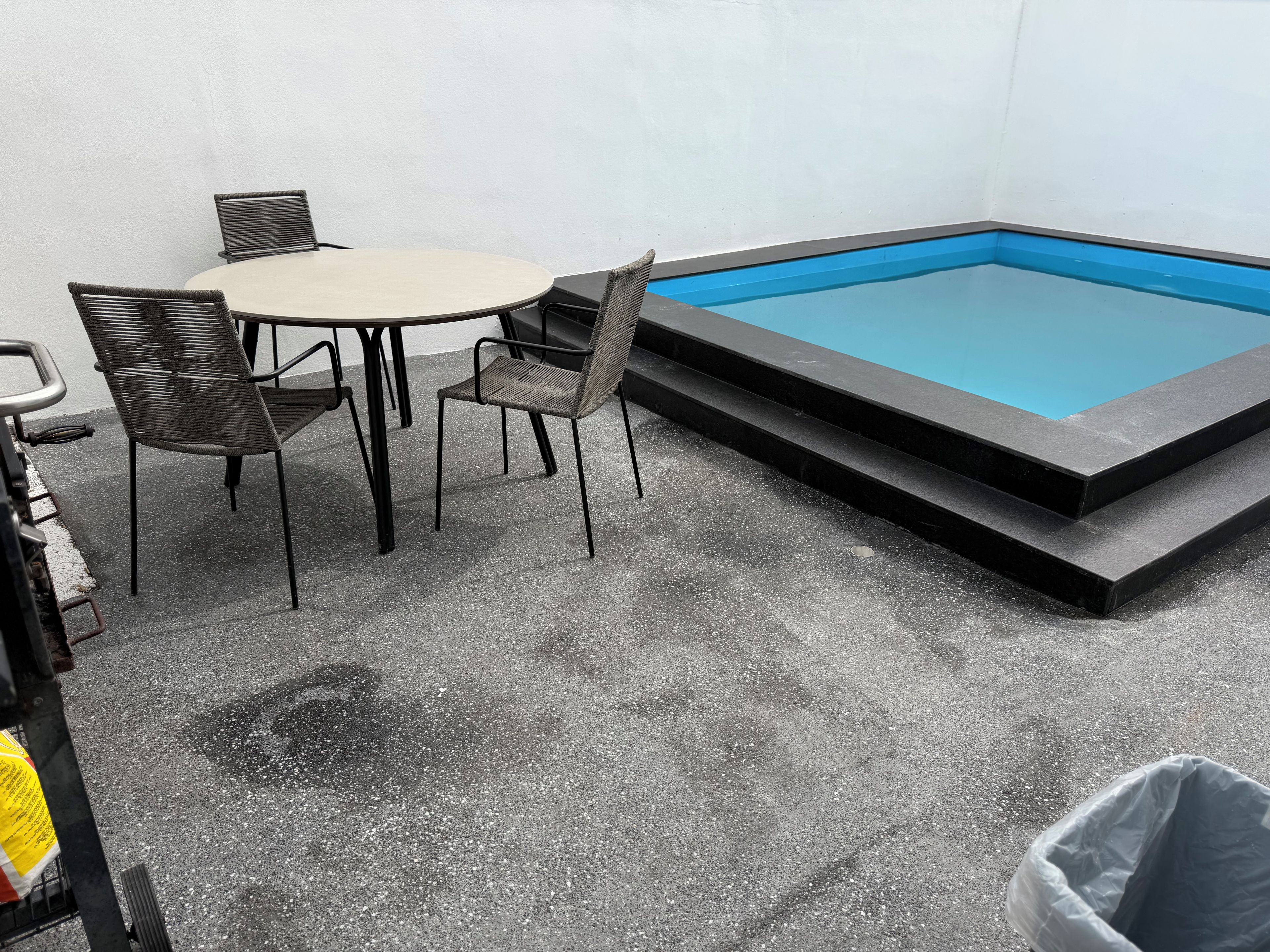 A small outdoor pool area with a table and chairs, a trash bin, and a concrete floor.