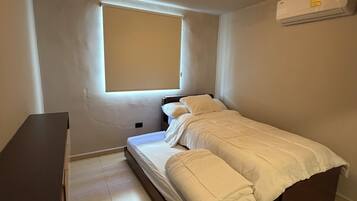 Ein kleines, ordentlich eingerichtetes Schlafzimmer mit einem Einzelbett, einem Nachttisch, einem Fenster mit Jalousien, einer Deckenleuchte und einer Klimaanlage.