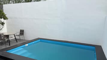 A rectangular swimming pool with a black edge, surrounded by a white wall and a small patio area with a table and chairs.