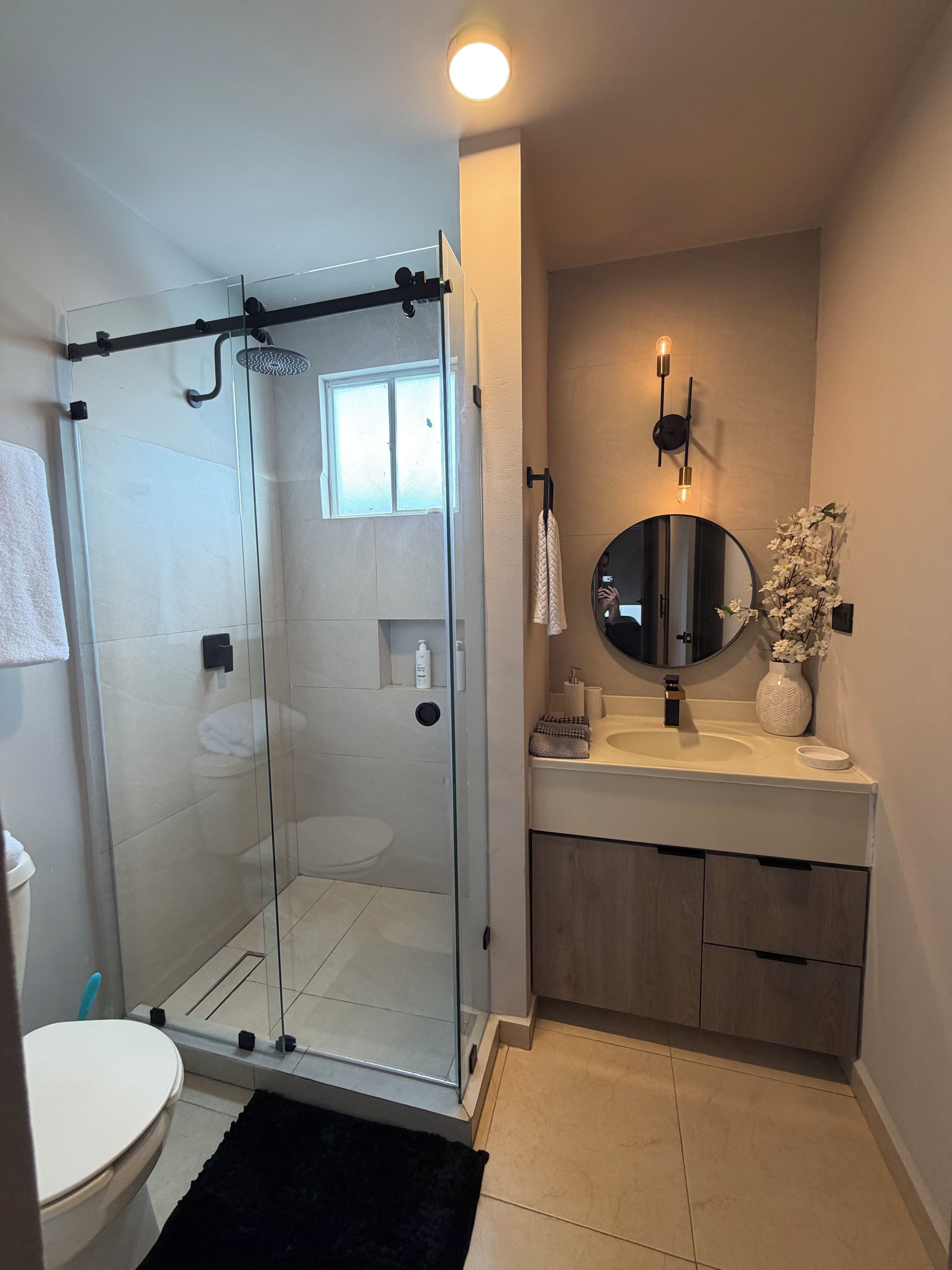 A modern bathroom with a glass-enclosed shower, a round mirror, a sink with a vase, and a toilet.