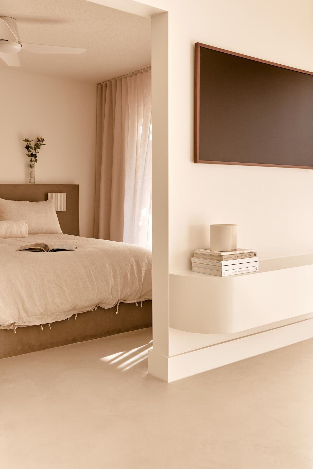 A minimalist bedroom with a bed, a bedside table with books, a vase with flowers, and a large mirror on the wall.