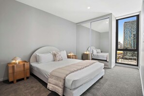 Un dormitorio moderno con una cama grande, mesitas de noche, un espejo y vistas a un paisaje urbano a través de un ventanal.