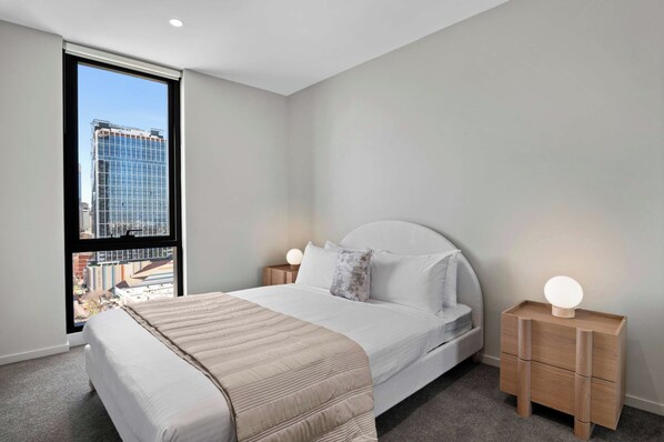 Habitación de hotel moderna con una cama grande, dos mesitas de noche con lámparas y vistas a un paisaje urbano a través de un ventanal.