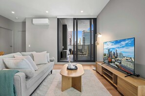 Una sala de estar moderna con un sofá gris, una mesa de centro, un televisor sobre un mueble de madera y vistas a la ciudad a través de un ventanal grande.