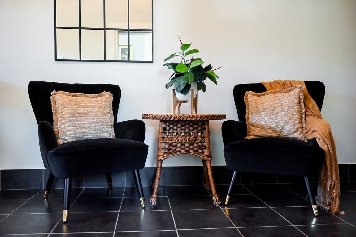Twee zwarte stoelen met geweven kussens, een klein rieten tafeltje met een potplant en een betegelde vloer.