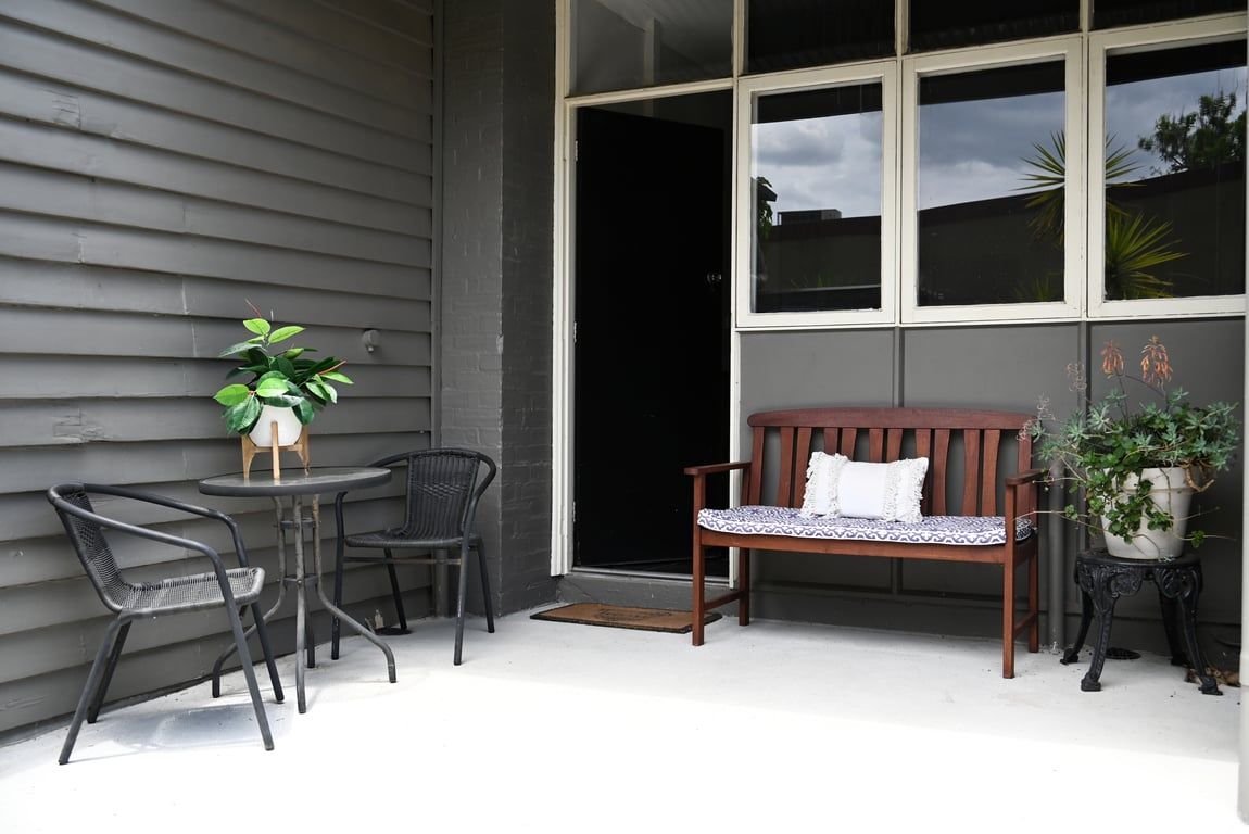Een terras met een houten bank, twee stoelen, een kleine tafel met een bloempot en een groot raam.