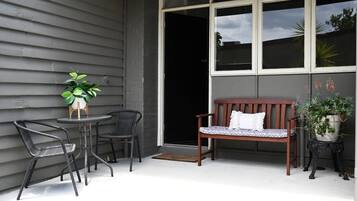 Een terras met een houten bank, twee stoelen, een kleine tafel met een bloempot en een groot raam.