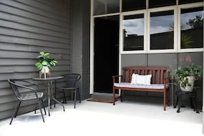 A patio with a wooden bench, two chairs, a small table with a potted plant, and a large window.