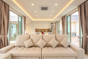 Ein modernes Wohnzimmer mit einem beigefarbenen Sofa, einer Küche im Hintergrund und großen Fenstern.