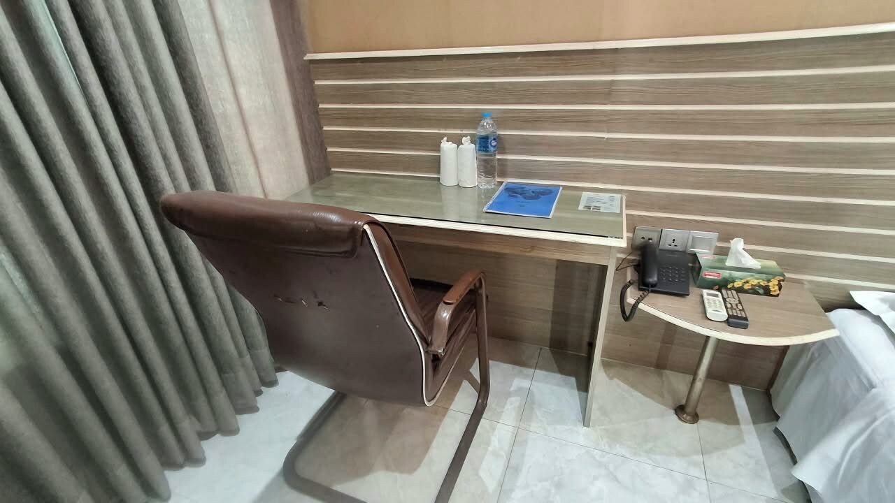 Ein Hotelzimmer mit Glastisch, braunem Ledersessel, kleinem runden Tisch mit Fernbedienung, Taschentuchbox und Wasserflasche.
