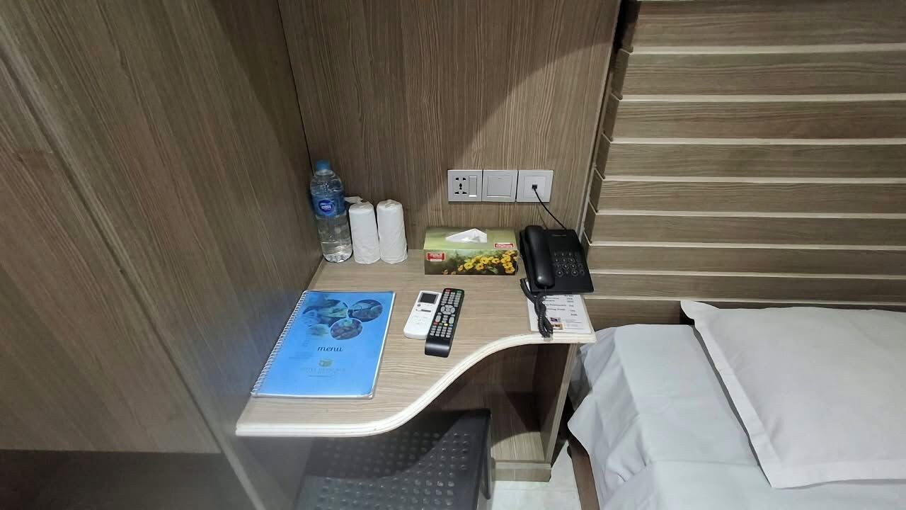 A compact hotel room with a wooden desk, a bed, a small table with a notebook, a remote control, a phone, and toiletries.