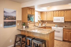 A kitchen with wooden cabinets, a granite countertop, a white stove, and a painting of a forest on the wall.