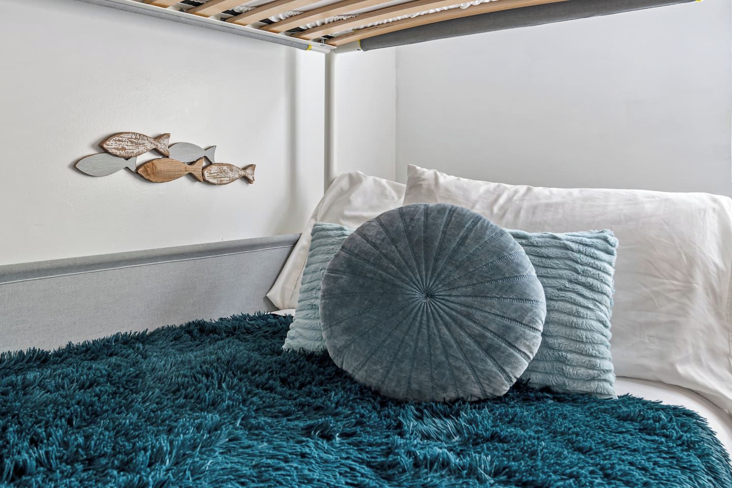 A bed with a teal furry rug, two pillows, and a wall with a wooden shelf holding decorative fish.