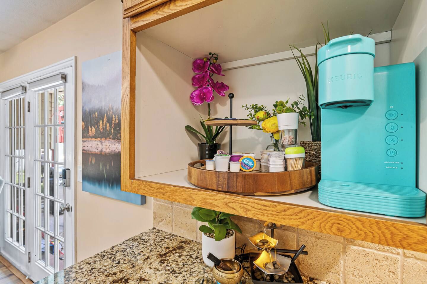 A kitchen with a coffee maker, a wooden shelf with various items, a painting, and a door.