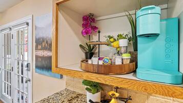 A kitchen with a coffee maker, a wooden shelf with various items, a painting, and a door.