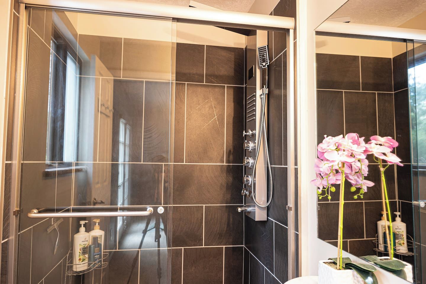 A modern bathroom with a glass shower enclosure, a handrail, and a mirror reflecting a pink orchid.