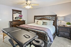 Een slaapkamer met een bed, nachtkastje, kaptafel, spiegel, plafondventilator en een massagetafel.