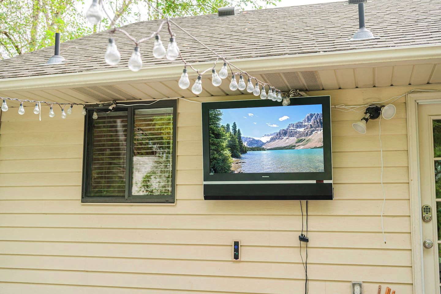 A house with a mounted television displaying a scenic landscape, a window with blinds, and a string of light bulbs.