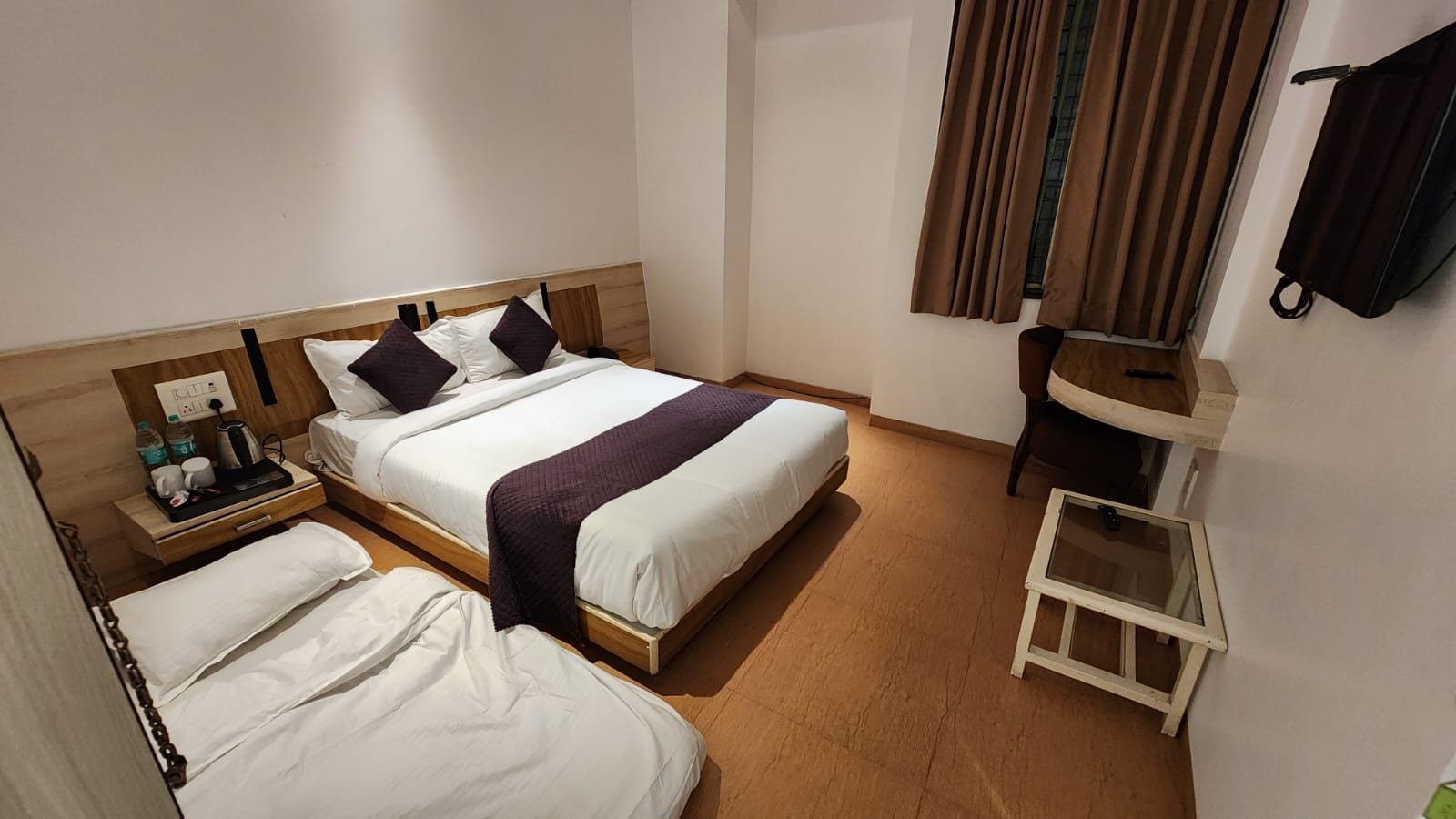 Een hotelkamer met twee bedden, een nachtkastje met een lamp, een klein tafeltje met een waterkoker en een raam met gordijnen.