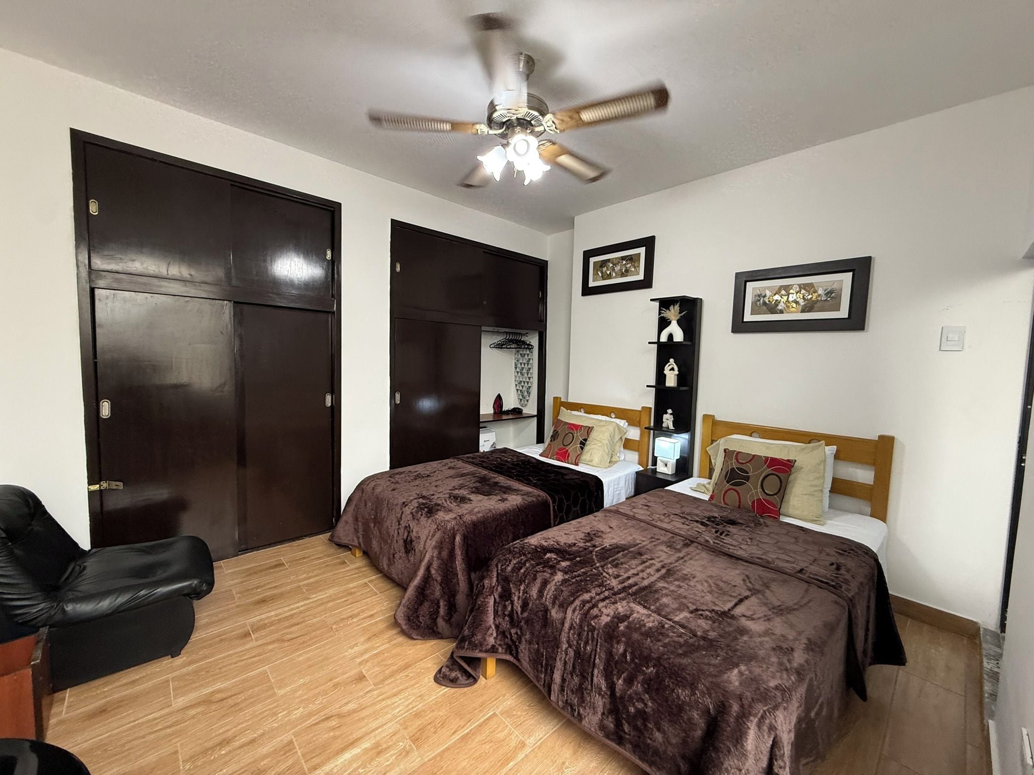 A bedroom with two beds, a ceiling fan, wooden flooring, and a black chair.