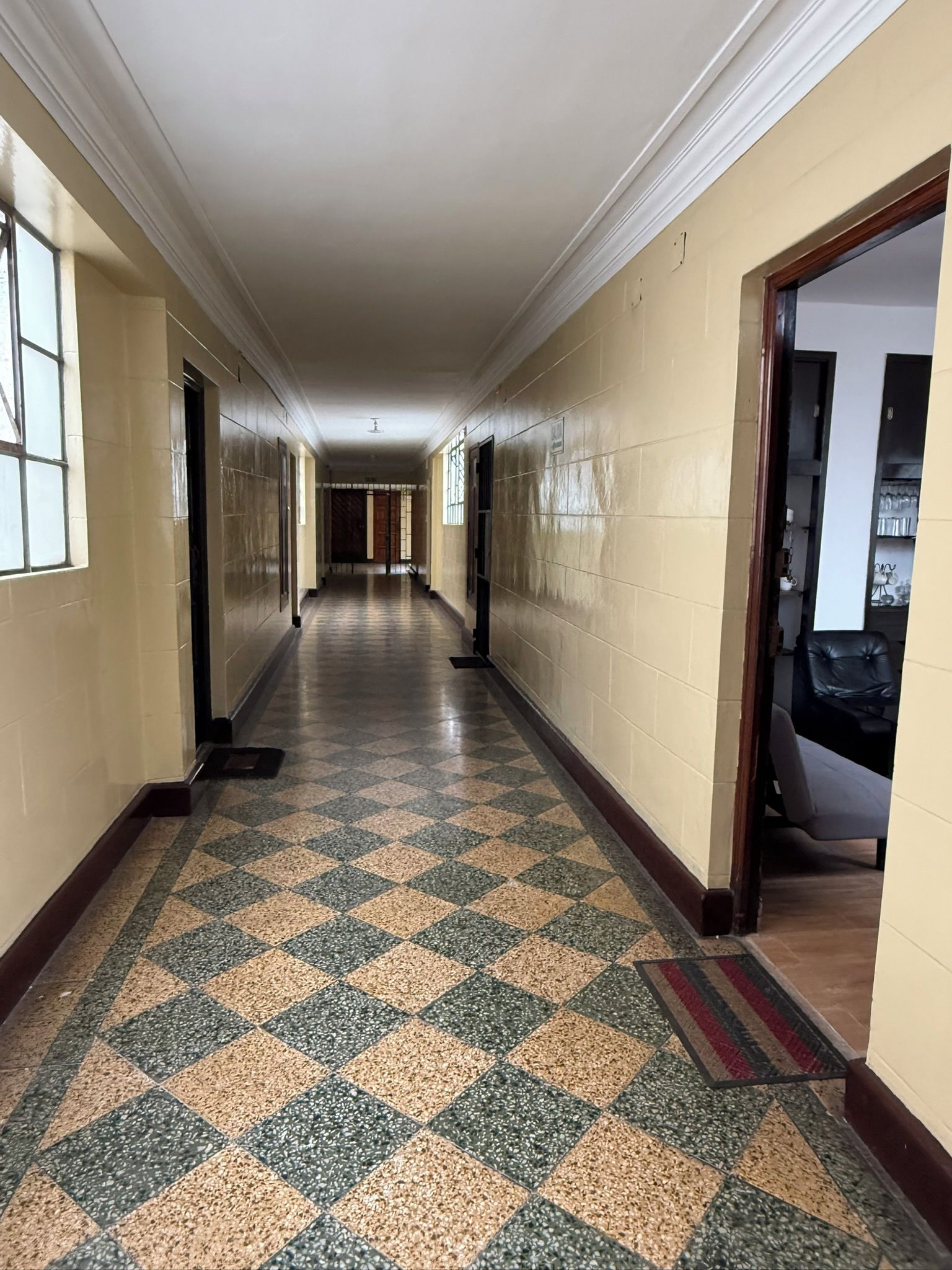 A long hallway with patterned flooring, doors on both sides, and a view into a room with a chair and a table.