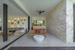 A modern interior with a stone wall, a ceiling fan, and a seating area with a white table and wicker chairs. - Villa Kadera (Pererenan)