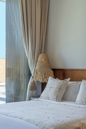A bed with a textured white cover, a woven hanging light fixture, and a view of the outdoors through a window with vertical blinds. - Villa Kadera (Pererenan)