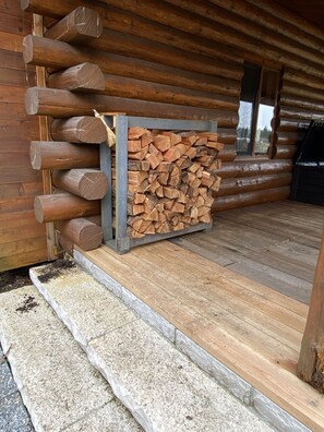 A wooden log cabin with a stack of firewood against the wall and a set of steps leading to a door.